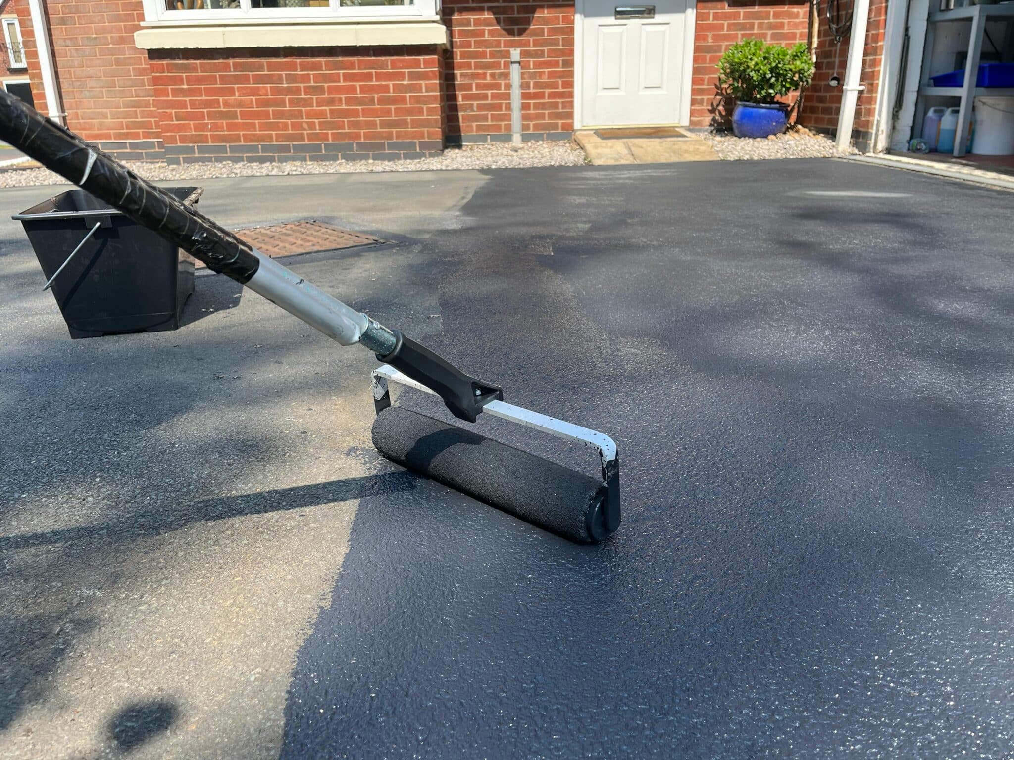 Wide roller applying black sealant across a residential driveway